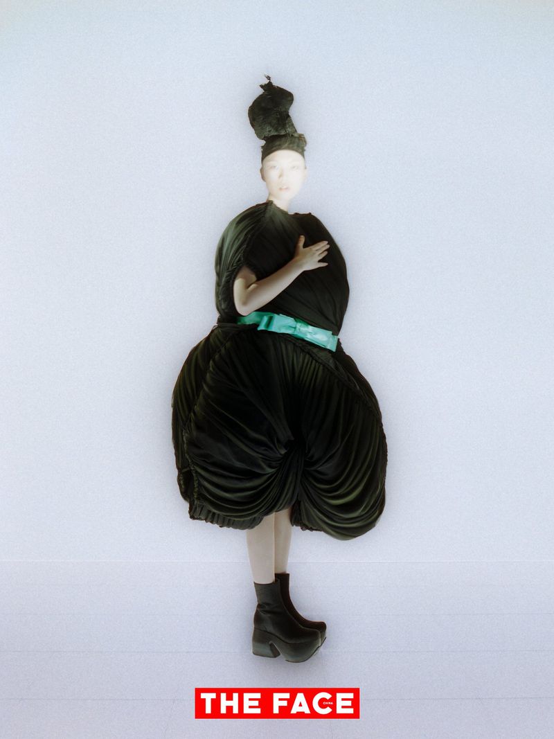 A person in a sculptural black avant-garde dress with a bright teal belt and high hair knot stands against a plain white background.