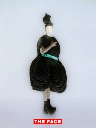 Model wearing a voluminous black pleated sculptural garment with a turquoise belt and tall headpiece against a white background.