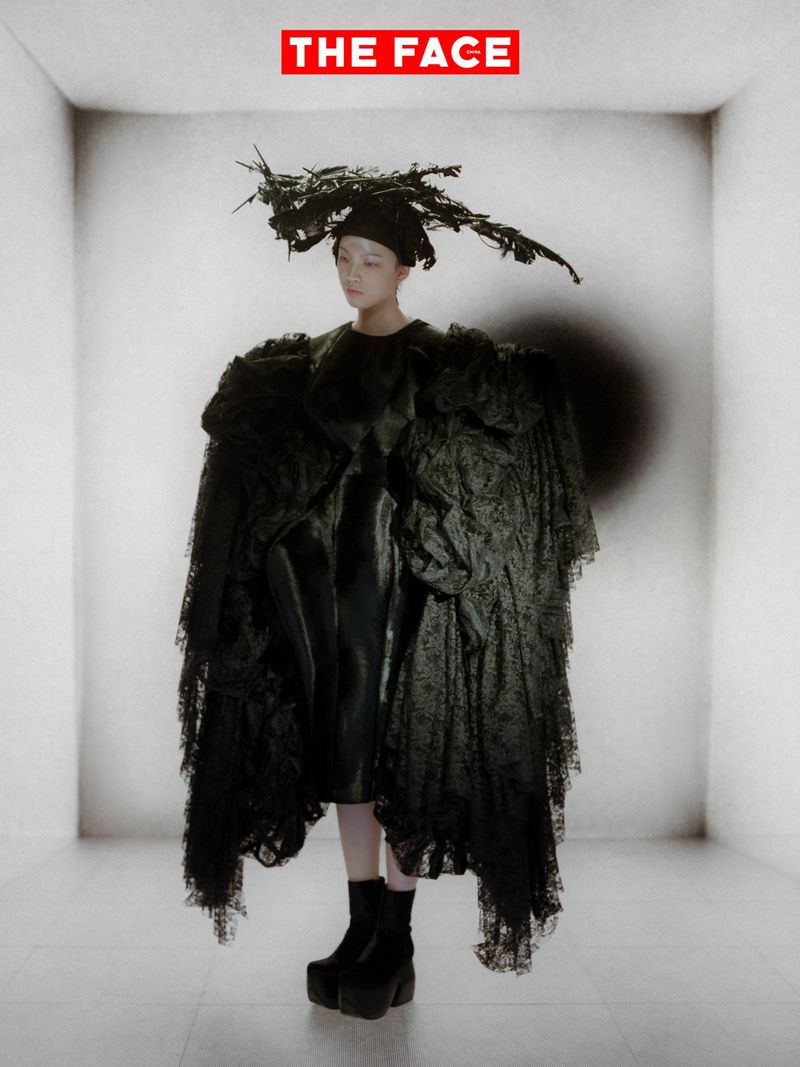 East Asian model in a voluminous black lace coat and sculptural twig headpiece posing in a white studio set.