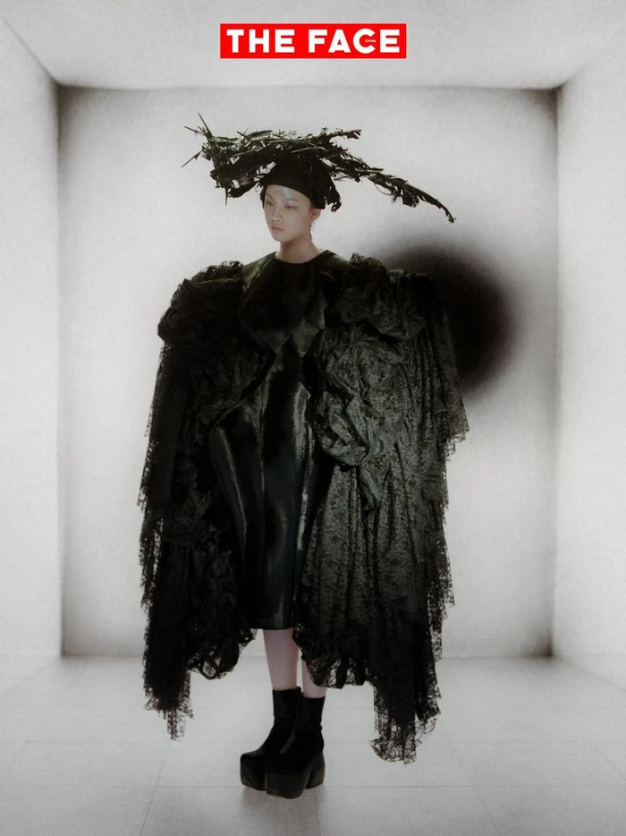 East Asian model in a voluminous black lace coat and sculptural twig headpiece posing in a white studio set.