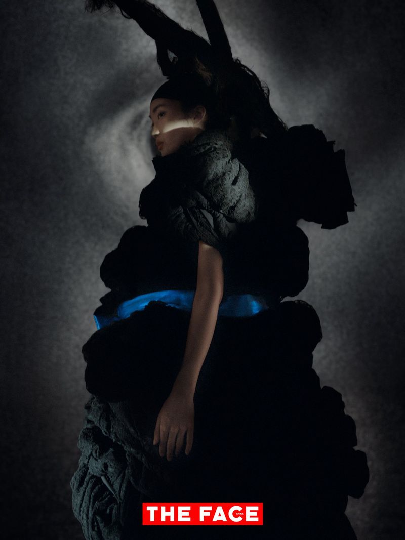 Model wearing a voluminous sculptural black garment with a vibrant blue waist detail against a dark textured backdrop.
