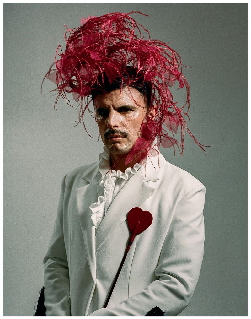 Rodrigo Cuevas in a white suit with ruffles and a large, elaborate red feather headpiece, holding a heart-shaped prop.
