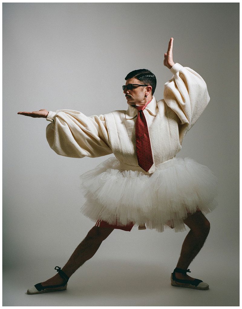 Rodrigo Cuevas posing dynamically in a white tutu, puffy long-sleeve top, sunglasses, and red tie against a studio backdrop.