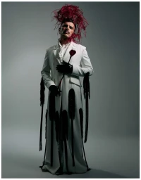 Rodrigo Cuevas posing in a full-length white coat with black fringe details and a large red feathered headdress against a grey studio backdrop.