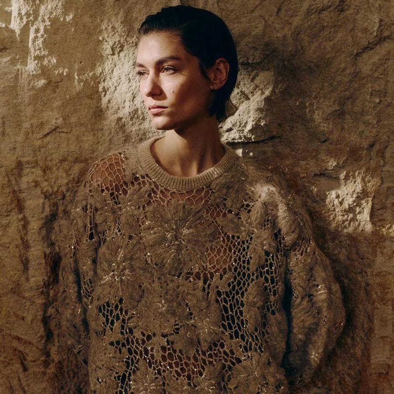 Model with slicked-back dark hair wearing an intricate beige openwork floral knit sweater against a textured stone wall.