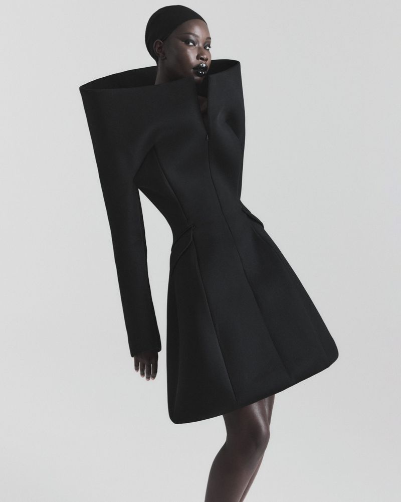 A model with dark skin wears a structural black mini coat with an oversized collar, standing against a neutral light gray background.