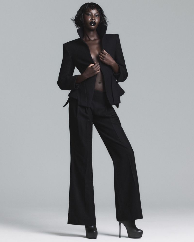 Model Adhel Bol posing in a structured black blazer with flared trousers and black platform heels against a light grey studio background.