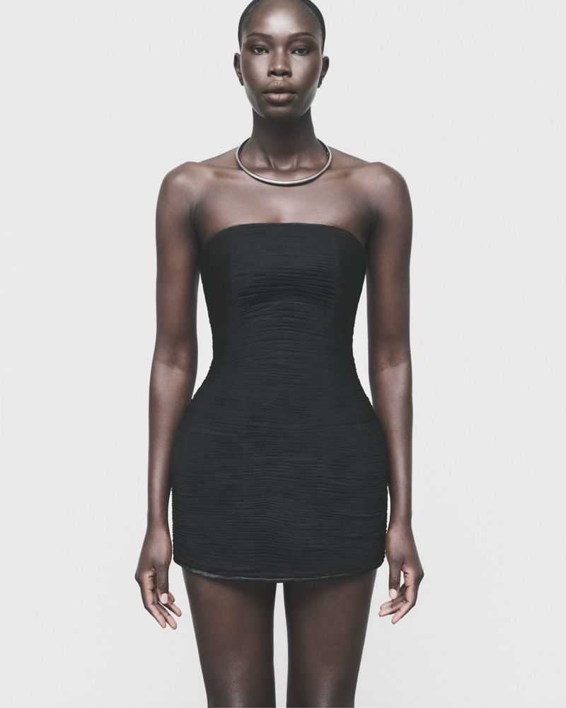 Model Adhel Bol wearing a strapless black mini dress and silver choker necklace against a neutral grey studio background.