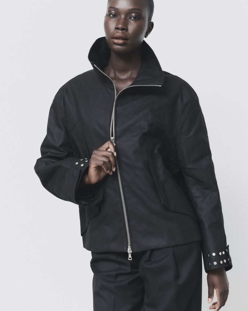 Model Adhel Bol wearing a high-collar black zip-up jacket with silver cuff studs against a pale backdrop.