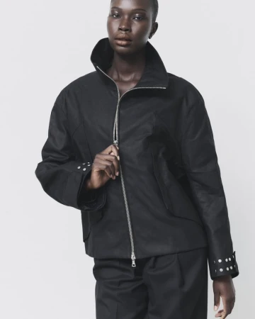 Model Adhel Bol wearing a high-collar black zip-up jacket with silver cuff studs against a pale backdrop.