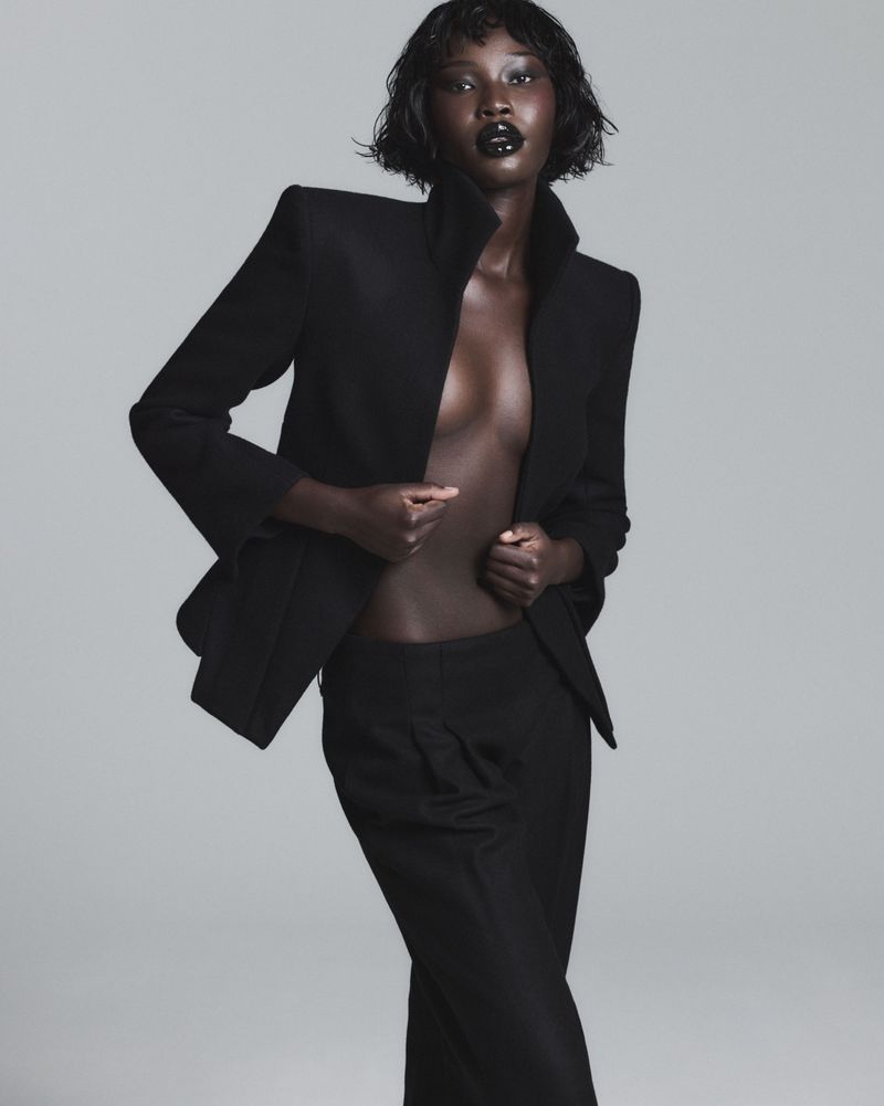 Model Adhel Bol wearing an open black tailored suit jacket and matching trousers against a smooth grey studio backdrop.