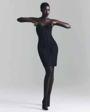 Model Adhel Bol stands in a strapless black corset mini dress, sheer black tights, and pointed heels against a grey studio backdrop.