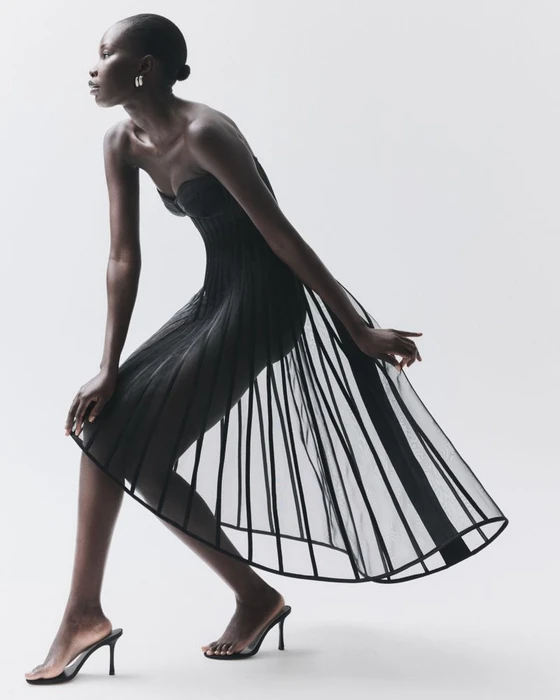 Model Adhel Bol wearing a sculptural black strapless sheer dress with heels against a plain white studio background.