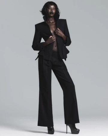 Model Adhel Bol posing in a structured black blazer with flared trousers and black platform heels against a light grey studio background.