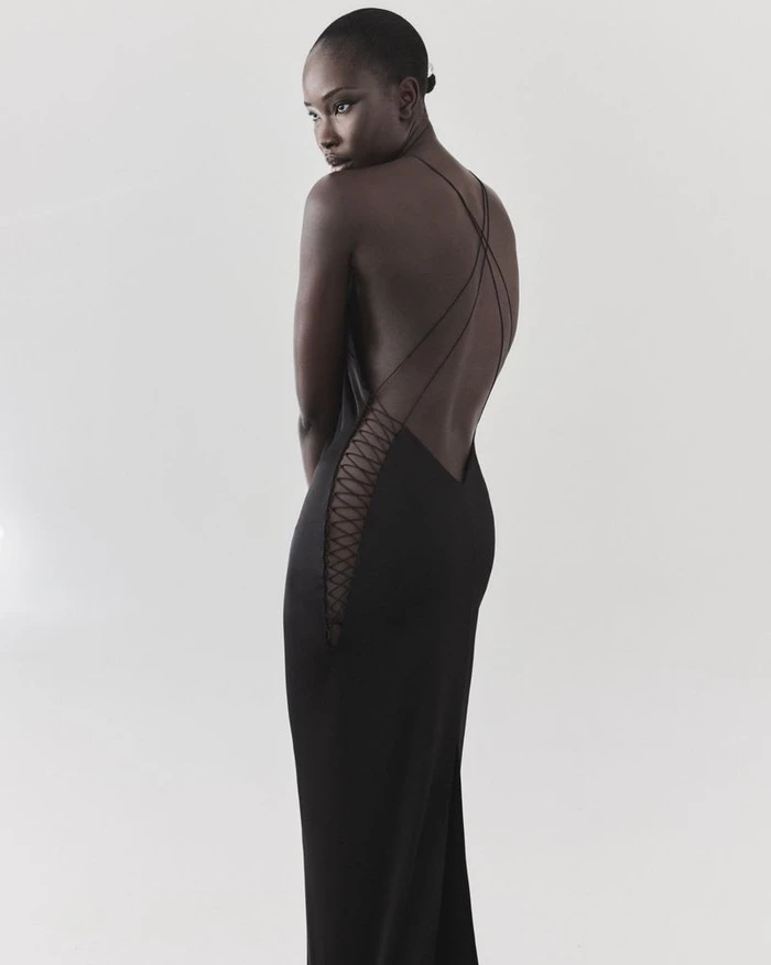 Model Adhel Bol wearing a black backless gown with crossed strap detailing against a white studio background.