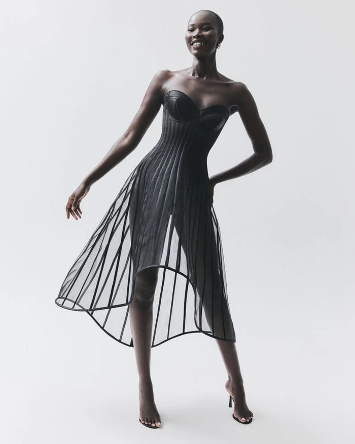 Model Adhel Bol smiling in a sheer black cage corset dress and black high-heel sandals against a white studio background.