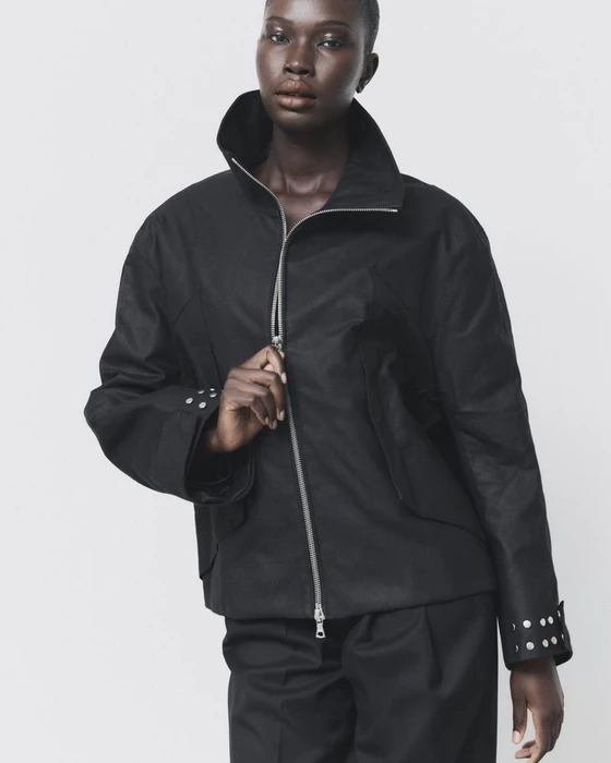 Model Adhel Bol wearing a high-collar black zip-up jacket with silver cuff studs against a pale backdrop.