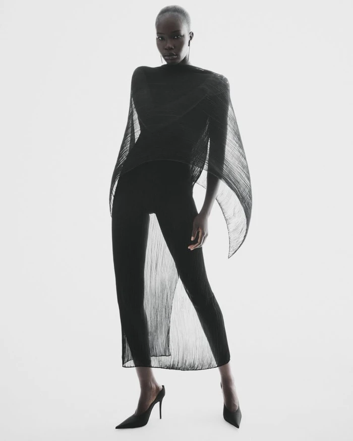 Model Adhel Bol posing in a black sheer draped capelet overlay, black leggings, and pointed stiletto heels against a white background.