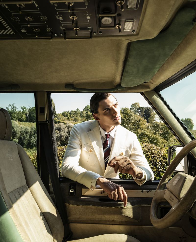 Man wearing a cream tailored blazer and striped necktie rests his arms on a vehicle door while looking upwards in natural light.
