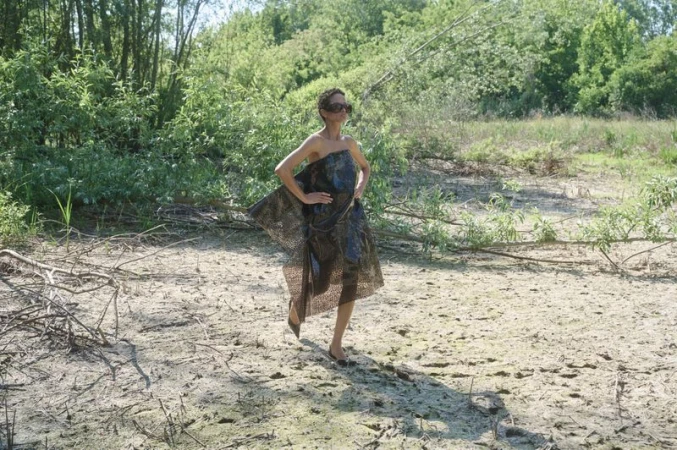 Model wearing dark sunglasses and a sheer patterned strapless dress walking across dry cracked ground against green trees.