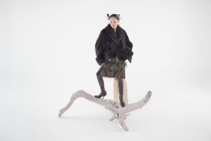 Model Xie Chaoyu posing atop bleached driftwood in a black textured coat and peacock feather skirt against a white studio background.