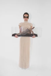 Model wearing dark sunglasses and a beige high-neck maxi dress holding a glass terrarium with driftwood against a white background