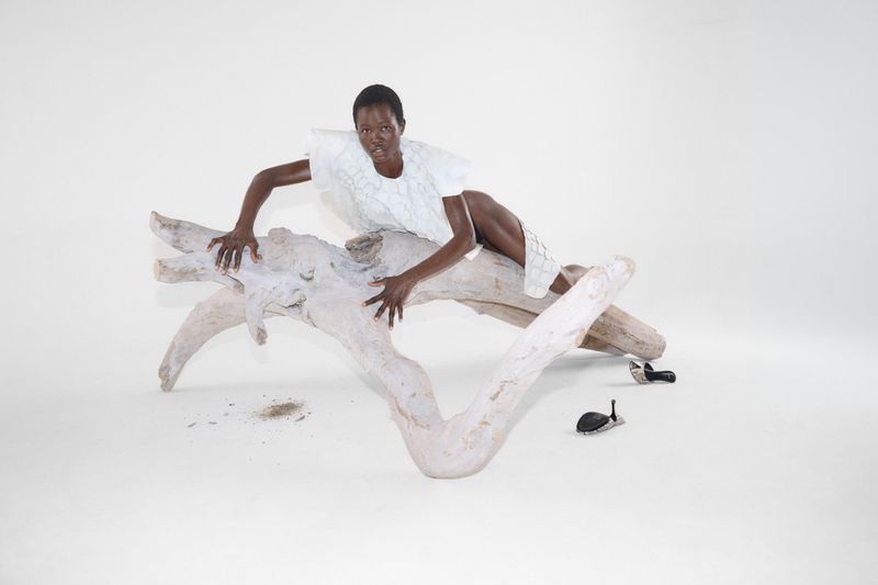Anyiel Majok poses across a large bleached driftwood branch in a white structured top against a white studio background.
