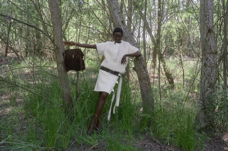 Model Anyiel Majok wearing a white textured top and matching skirt holding a brown handbag between forest trees.