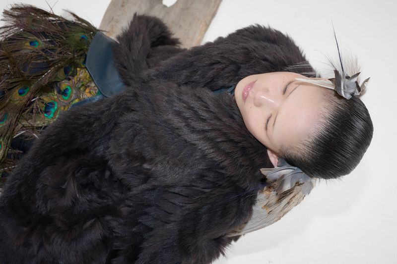 East Asian model with sleek hair and feather accessories wearing a dark textured fur garment against a white background.
