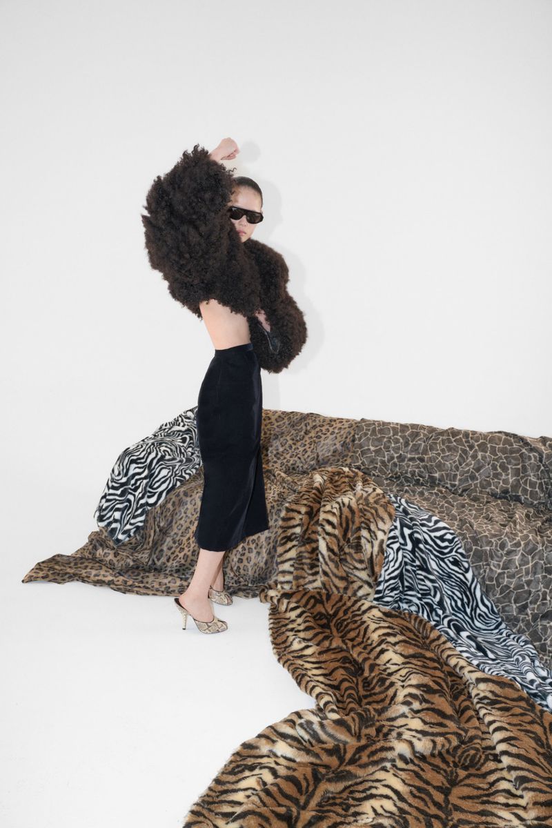 Model wearing dark sunglasses, an oversized brown fur jacket, and a black skirt standing beside draped animal print fabrics in a studio.