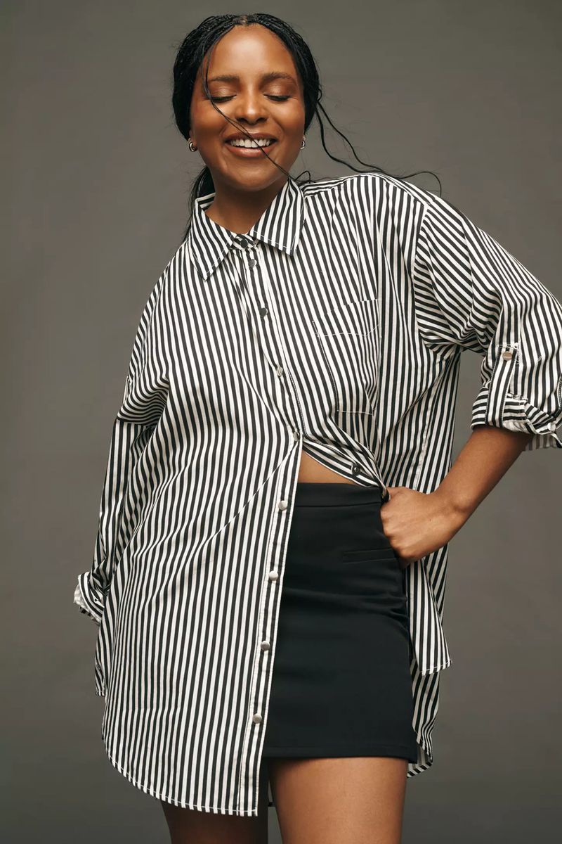 A smiling African-American woman poses in a black and white striped shirt and black skirt against a grey background in a medium shot.