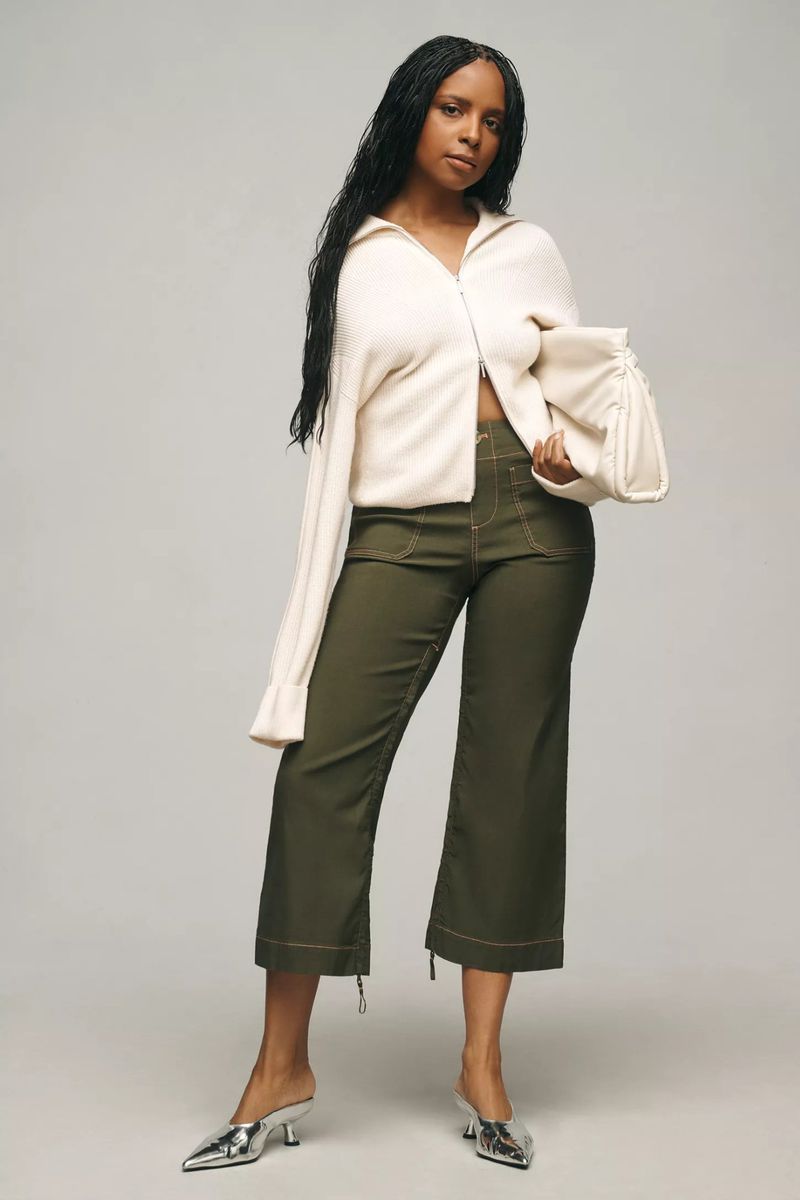 A Black woman in a cream sweater, olive pants, and silver heels poses in a full shot against a neutral studio backdrop.