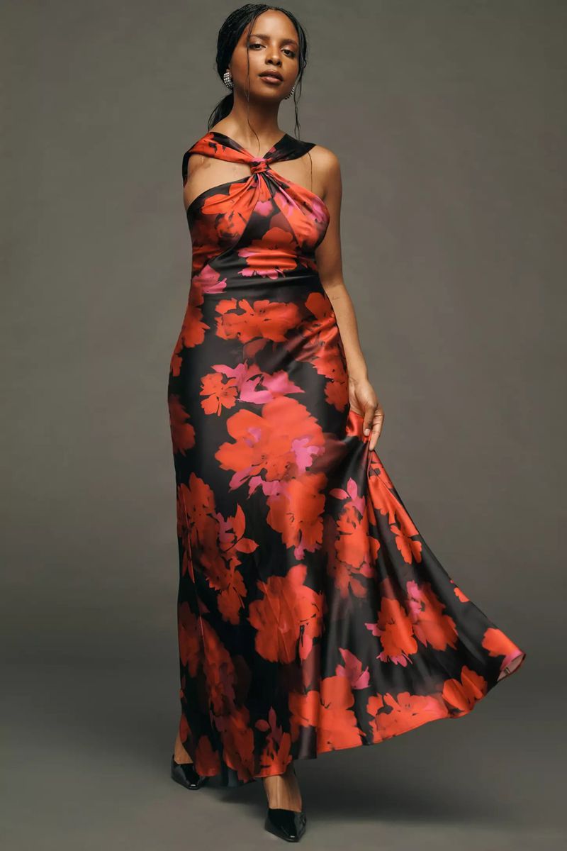A woman in a floral gown poses in a studio. The one-shoulder dress is black with red flower pattern, and she wears black shoes.