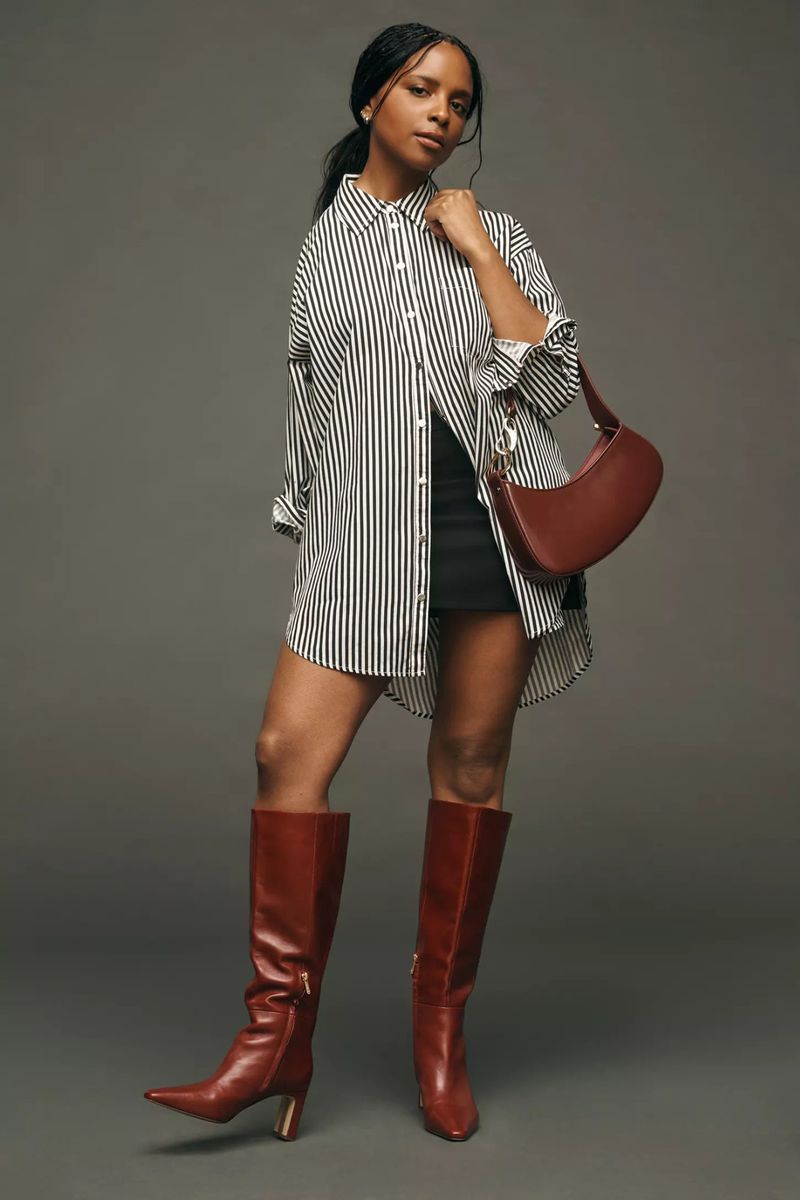 A confident Black woman posing in a striped shirt, black skirt, and brown boots with a matching purse against a grey background.