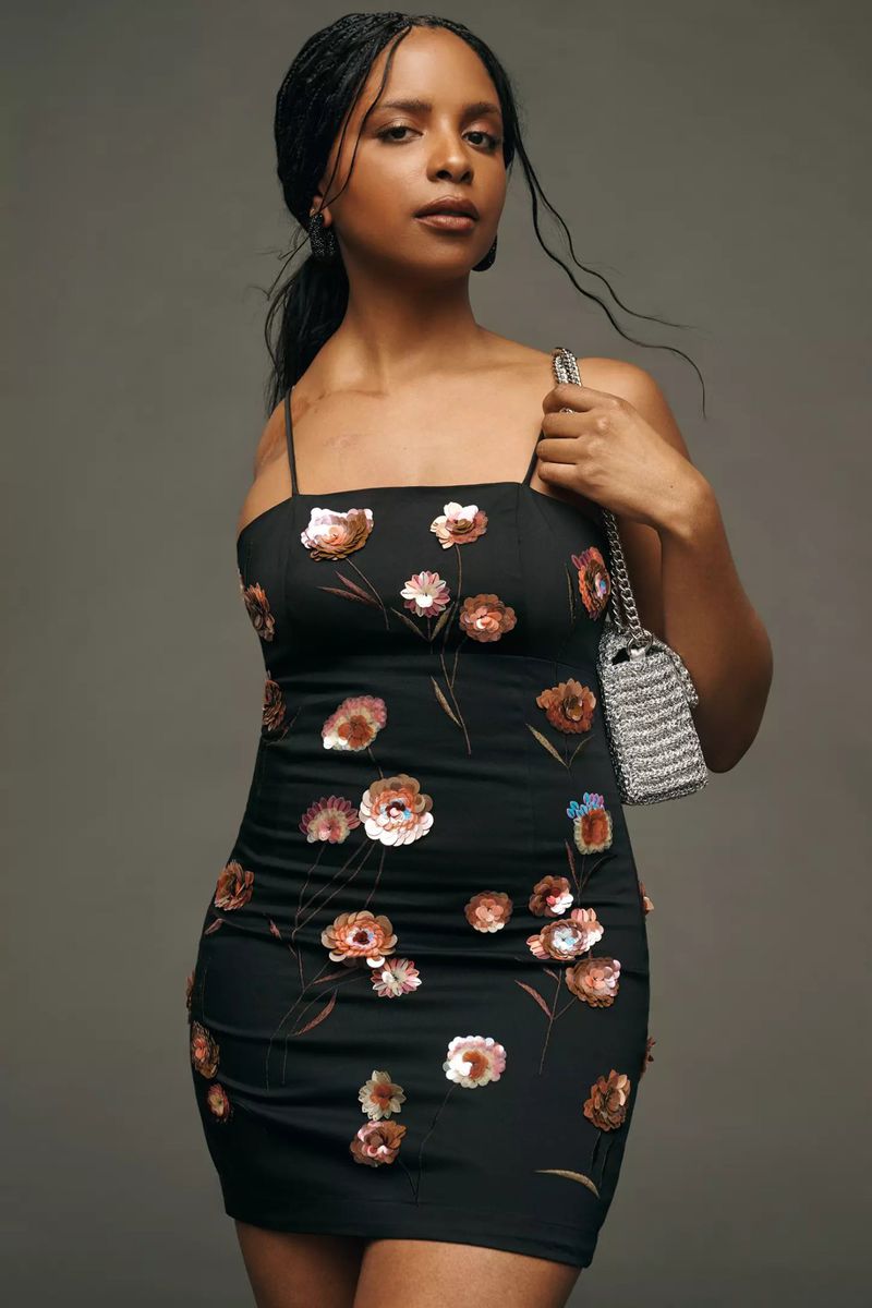 Adult female poses indoors wearing a black floral dress and a silver purse over her shoulder in a studio setting on gray backdrop.