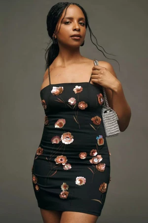 Adult female poses indoors wearing a black floral dress and a silver purse over her shoulder in a studio setting on gray backdrop.