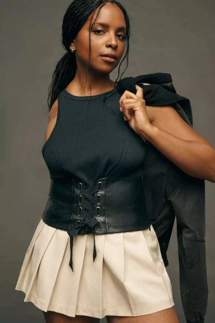 Portrait of a woman with braided hair, wearing a black top, leather corset, and pleated skirt, holding a black jacket on her shoulder.
