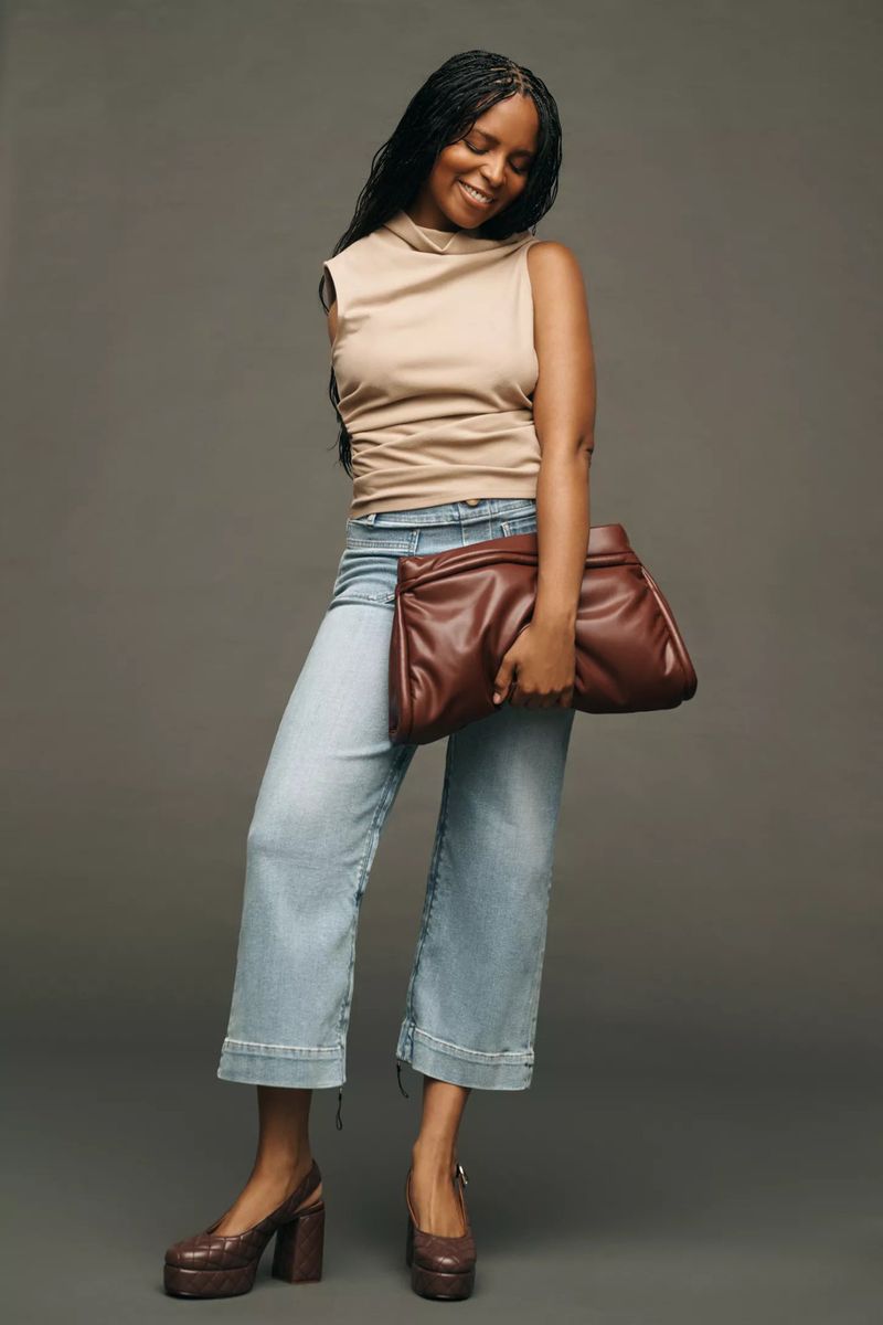 Woman in beige top and light denim jeans posing with a large brown leather clutch against a grey studio backdrop.