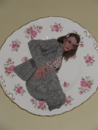 Fashion model in a grey textured suit composited inside a vintage porcelain plate with pink floral patterns on a plain wall.