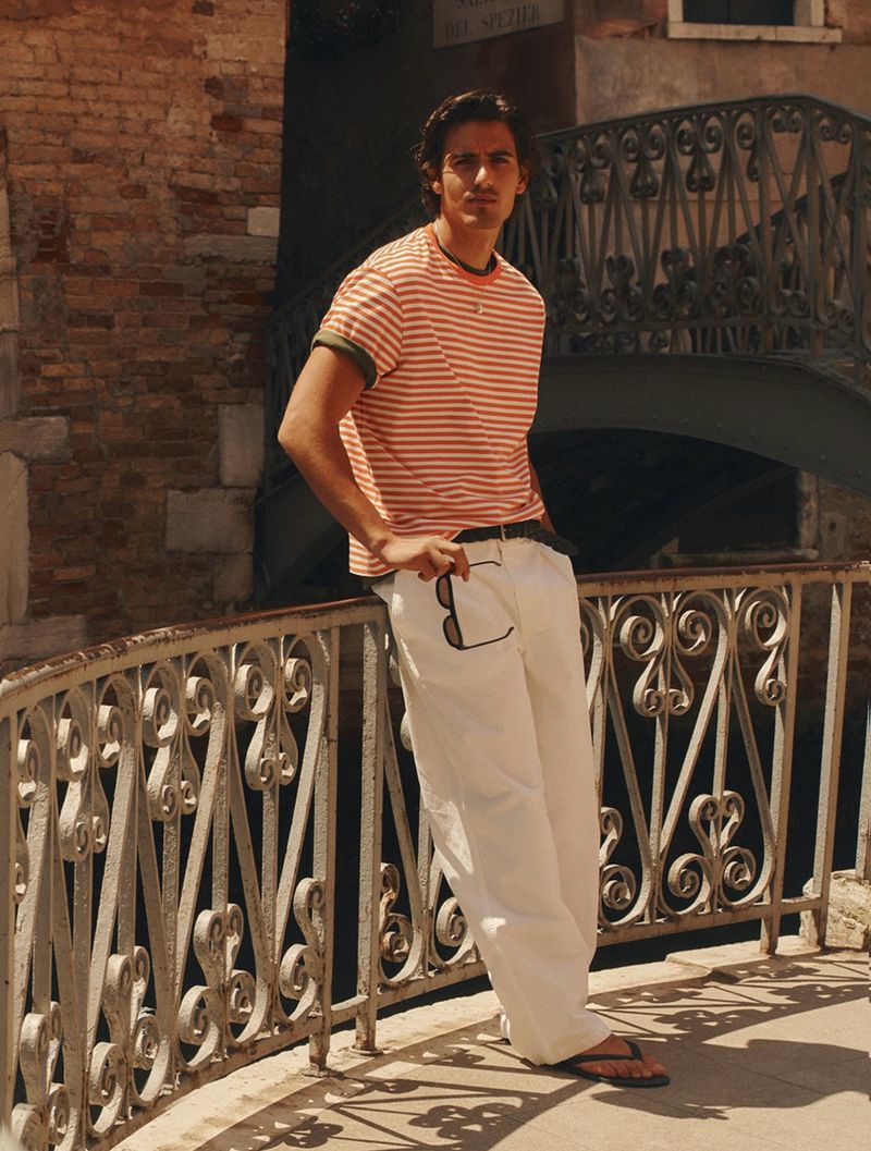Man wearing a striped t-shirt and white trousers leaning against a metal bridge railing in Venice, Italy.
