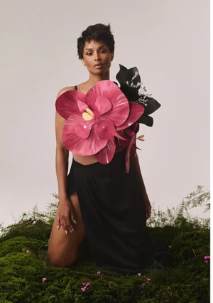 Ciara kneeling on a moss patch wearing an oversized sculptural pink orchid top and a black high-slit skirt in a studio setting.