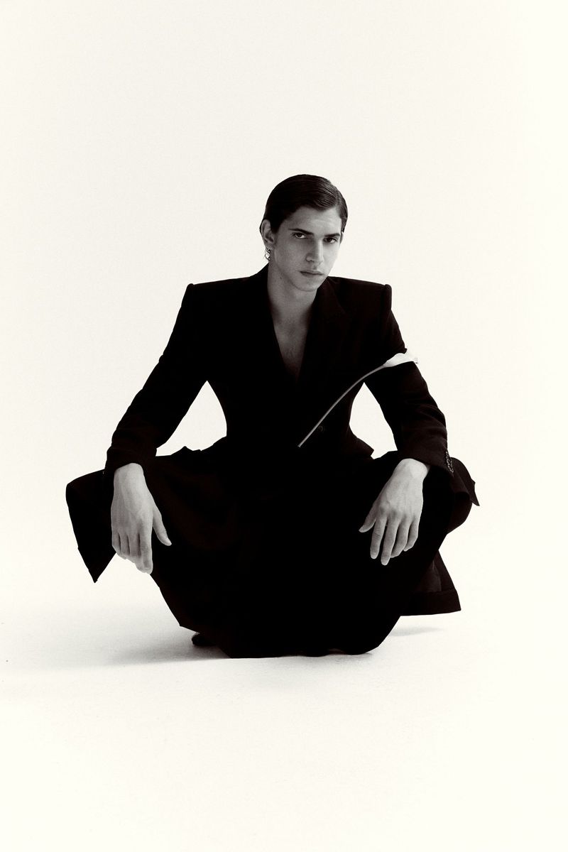 Model Hedi Ben Tekaya crouching in a tailored black suit against an unbroken white studio backdrop.
