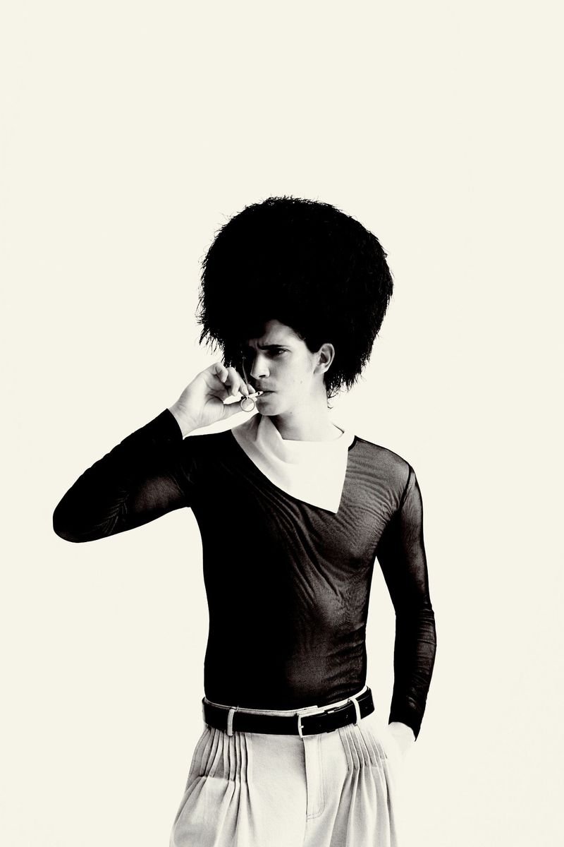 A person with a large afro hairstyle wearing a black sheer long-sleeve top and light-colored trousers poses against a plain background.