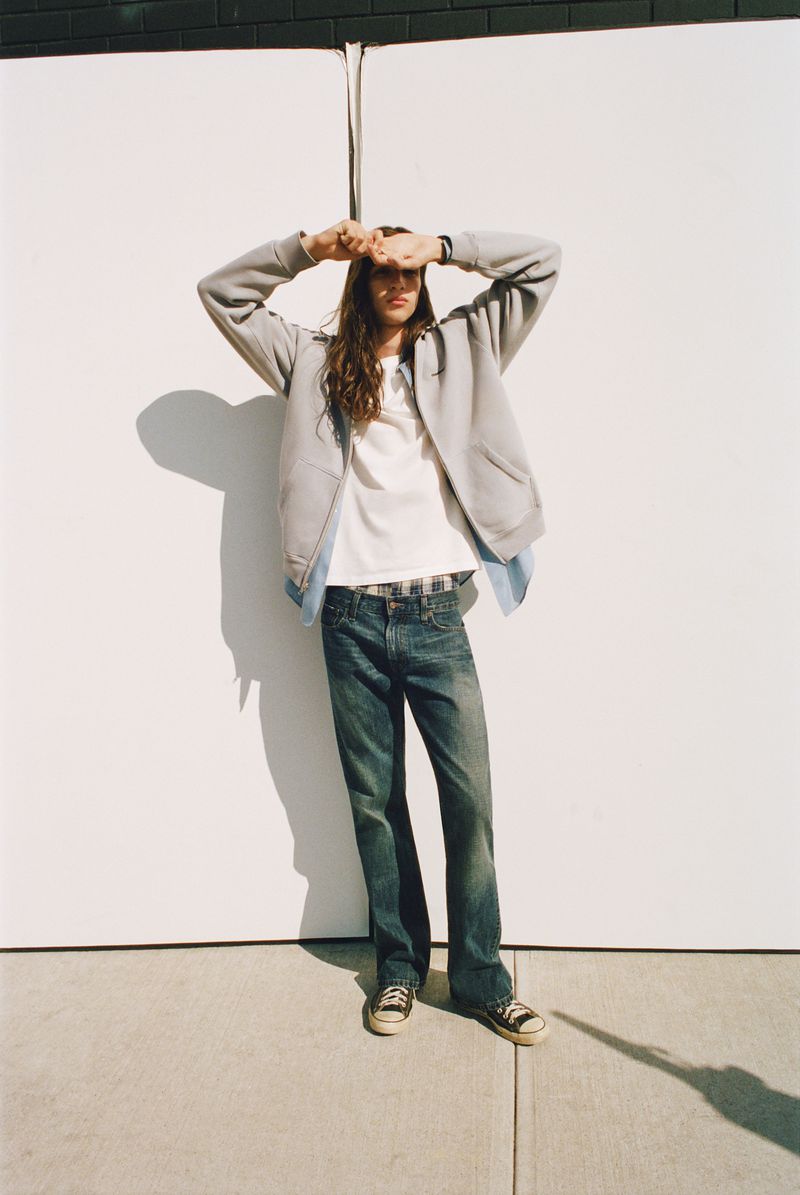 Model posing in a grey zip hoodie, white t-shirt, and blue denim jeans against a white wall in direct sunlight.