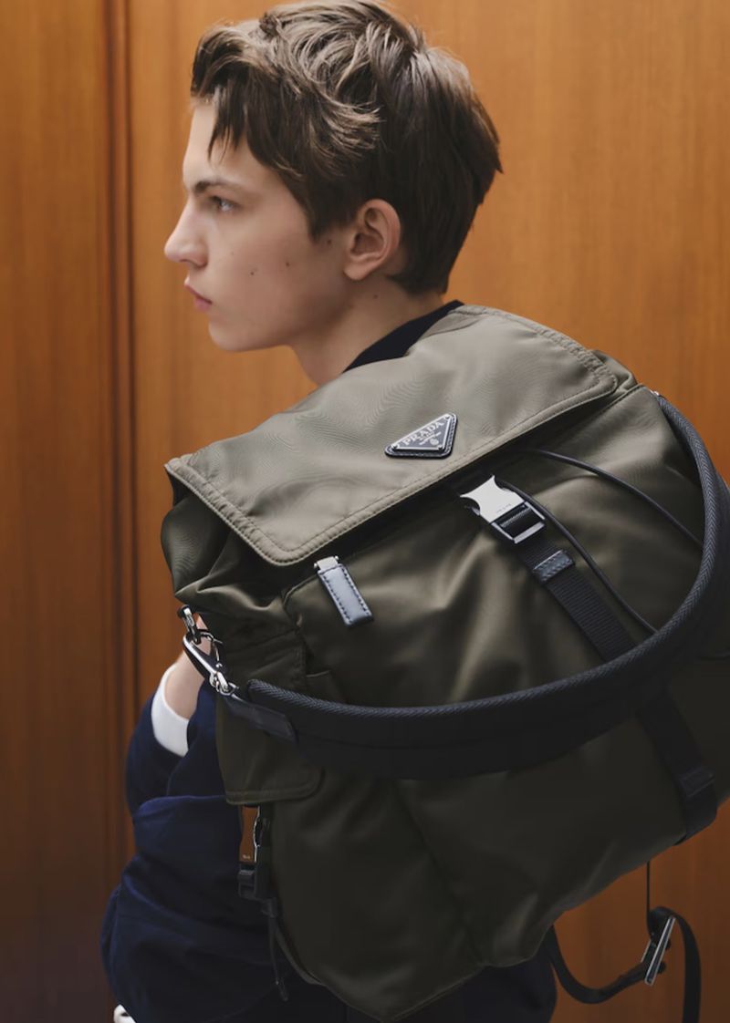 Young man with short brown hair seen in profile, wearing a dark jacket and carrying an olive green Prada nylon backpack.
