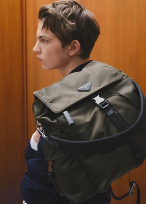 Model Akim Bats wearing an olive green Prada nylon backpack over his shoulder against a warm wood panel background.