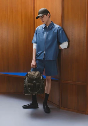 Male model in a blue zip-up short-sleeve shirt and matching shorts holding a dark quilted backpack against wood-paneled walls.