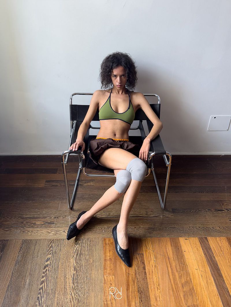 Young adult female poses in a green top, brown skirt, black ballet shoes, and gray knee pads while seated in a chrome chair.