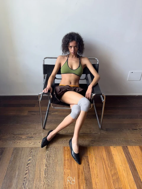 Young adult female poses in a green top, brown skirt, black ballet shoes, and gray knee pads while seated in a chrome chair.