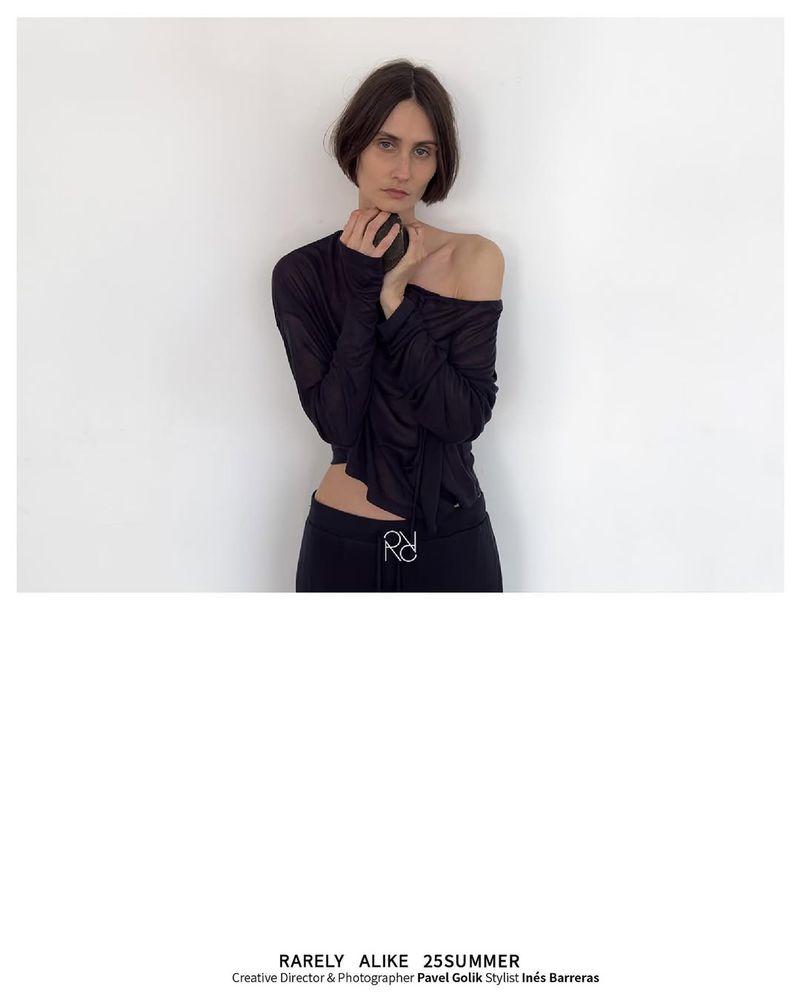 A woman with dark hair is posing in a black top and pants against a white wall in a fashion editorial studio setting.